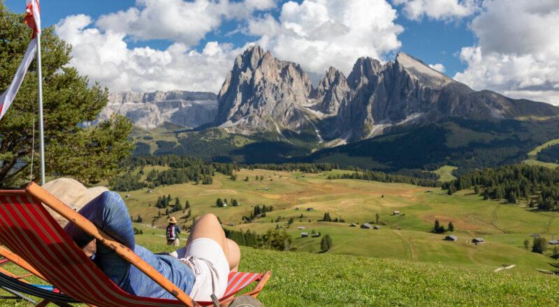 Frau liegt entspannt in einer Liege auf einer Wiese, nahe einer Ferienhütte in den Bergen